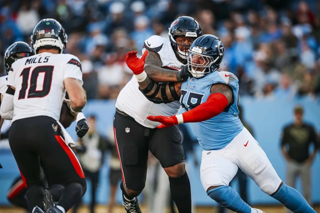 A football player in a blue uniform is tackling a player in a red and black uniform, with another player in a white uniform visible in the background. The scene depicts an intense moment during an NFL game.
