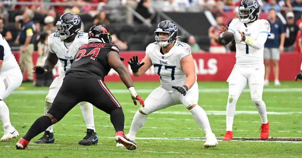 Tennessee Titans players on the field during a football game. A quarterback is preparing to throw a pass while offensive linemen block a defender in a black uniform.