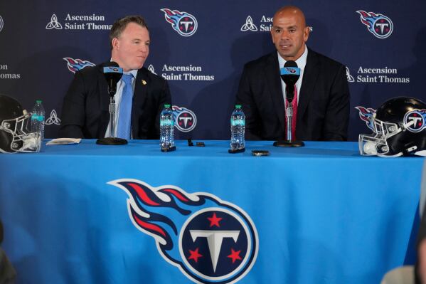 Two men seated at a table during a press conference, with a backdrop featuring the Tennessee Titans logo. The table is adorned with bottled water and microphones.